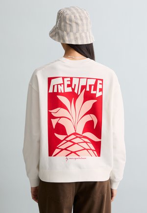 Osoba ubrana w białą bluzę z dużym czerwonym motywem ananasa oraz stylizowanym napisem „PINEAPPLE” na plecach, w połączeniu z czapką typu bucket w kratę.