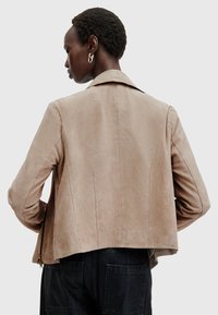 Giacca in suede beige con colletto strutturato, vestibilità su misura e dettagli di cucitura visibili. Tasca con zip sul lato, abbinata a pantaloni scuri.