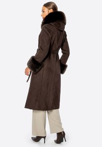 Brązowy zamszowy płaszcz z futrzanym kapturem i mankietami, z paskiem w talii oraz długą sylwetką. Modelka nosi beżowe spodnie i brązowe botki na obcasie.