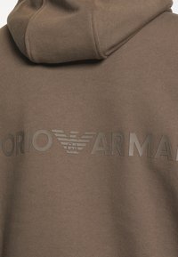 Brown hoodie with a textured finish, featuring a raised logo in dark color reading "Emporio Armani" and a stylized eagle emblem.
