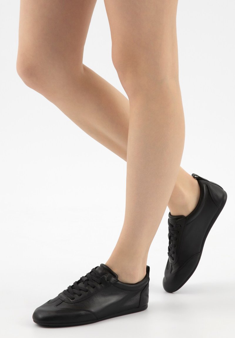 Beine, die niedrig geschnittene schwarze Leder-Sneaker mit Schnürsenkeln tragen, auf einem weißen Hintergrund positioniert, wobei ein Fuß leicht hinter dem anderen angehoben ist.