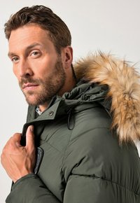Homme aux cheveux bruns courts et à la barbe, portant une veste en doudoune verte avec capuche en fourrure, ajustant son col, regardant l'appareil photo sur un fond neutre.
