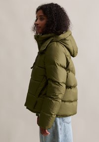 Oliwkowa kurtka puchowa z kapturem, pikowana tekstura, zamek błyskawiczny z przodu i kieszenie boczne. W zestawie z jasnoniebieskimi dżinsami.