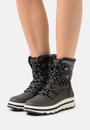 Schwarze Winterstiefel mit Pelzbesatz, gestreiften Schnürsenkeln und dicken weißen Sohlen, getragen an nackten Beinen vor einem schlichten Hintergrund.