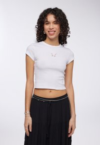 T-shirt corto a coste bianco con ricamo di una farfalla sul petto. Abbinato a una gonna nera fluida con una cintura decorativa.