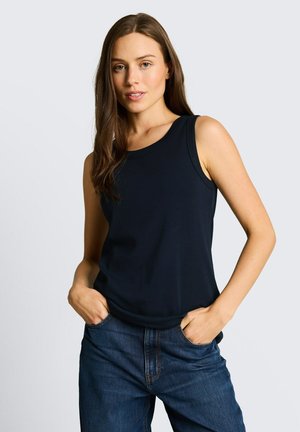 Jonge vrouw met lang bruin haar, een mouwloos zwart topje en blauwe spijkerbroek, handen in de zakken, tegen een effen lichte achtergrond.