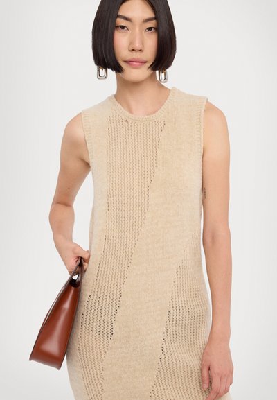 Beige krāsas adīta bezpiedurkņu kleita ar strukturētām rakstām un atvērtu šuvju detaļām. Modelis tur brūnu ādas rokassomu.