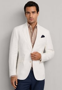 Mand iført hvid blazer med sort lommetørklæde, beige skjorte med knapper og marineblå bukser, stående mod en ensfarvet grå baggrund.