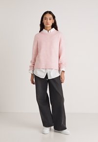 Ροζ πλεκτό πουλόβερ με στρογγυλή λαιμόκοψη, συνδυασμένο με λευκό πουκάμισο με κουμπιά και χαλαρά μαύρα τζιν, ολοκληρωμένο με λευκά αθλητικά παπούτσια.