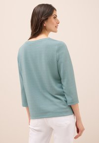 Top teal a maniche tre quarti con un motivo testurizzato, scollatura rotonda e tessuto morbido, abbinato a pantaloni bianchi, visto da dietro.