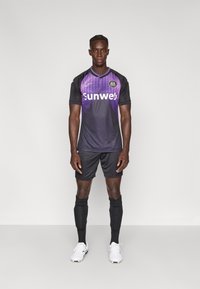 Μαύρη και μωβ φανέλα ποδοσφαίρου με στρογγυλή λαιμόκοψη και κοντά μανίκια, με λογότυπο "Sunweb". Συνδυάζεται με μαύρο σορτς και κάλτσες, λευκά αθλητικά παπούτσια.