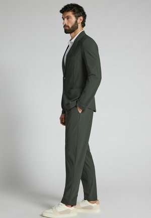Homme barbu portant un costume vert foncé sur mesure et des baskets blanches, debout de profil sur un fond clair uni.