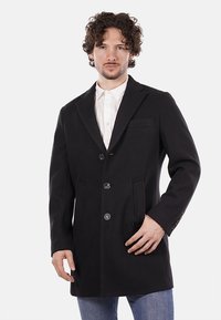 Abrigo de mezcla de lana negro con solapas de muesca, tres botones, dos bolsillos frontales y tela texturizada. Llevado sobre una camisa blanca y jeans.