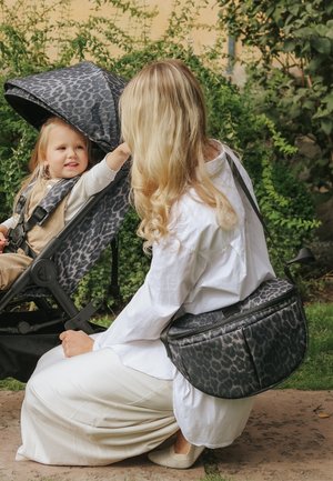 Donna con capelli biondi lunghi inginocchiata all'aperto, di fronte a un bambino piccolo sorridente nel passeggino con stampa leopardata, entrambi vestiti con abiti dai toni neutri.