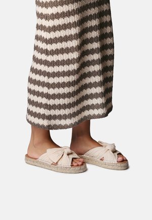 Fødder iført beige flade sandaler med åbne tæer og store snoede stofsløjfer, stående på en hvid og brunstribet strikket nederdel.