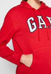 Sudadera roja hecha de tela suave con un bolsillo frontal. Presenta un gran logo bordado de 'GAP' en azul marino y blanco en el pecho.