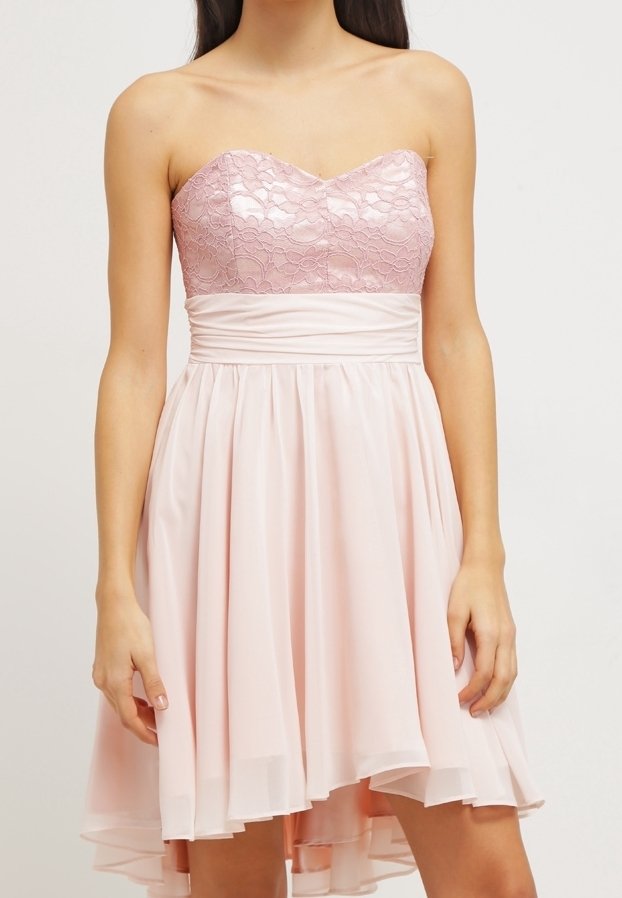 Femme portant une robe rose pâle sans bretelles avec un corsage en dentelle et une jupe fluide, debout devant un fond uni.