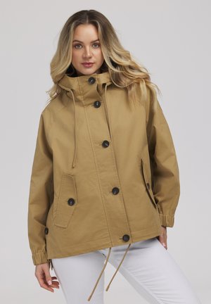 Kobieta o blond włosach, ubrana w beżową zapinaną na guziki kurtkę z wysokim kołnierzem i białe spodnie, na jednolitym tle.