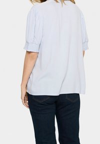 Blusa azzurro chiaro a maniche corte con maniche a sbuffo e dettagli smock sui polsini; vestibilità comoda; indossata con jeans in denim scuro.