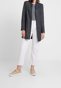 Femme portant un long manteau gris foncé, un haut gris, un jean blanc raccourci et des baskets beiges, debout devant un fond clair uni.