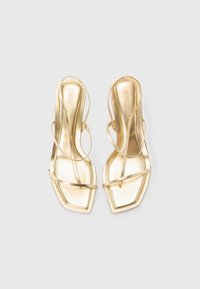 Tacchi slingback dorati con design a punta quadrata, caratterizzati da sottili cinturini incrociati e una texture metallica liscia. Design sovrapposto piatto.