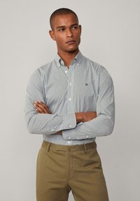 Camicia a righe blu e bianca, con logo verde, abbinata a pantaloni marroni. Tessuto morbido, colletto classico e maniche lunghe.