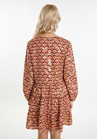 Rochie roșie cu un model de evantai floral în auriu și crem. Mâneci lungi, decolteu rotund și fustă în straturi cu o croială relaxată. Siluetă largă.