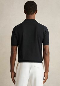 Tricou polo scurt cu mânecă, de culoare neagră, fabricat dintr-un material moale, având un guler clasic și un tiv ribben. Vederea din spate evidențiază un croi lejer.