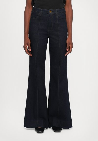 Jeans larges taille haute bleu foncé avec des coutures avant portés par une personne se tenant contre un fond blanc uni.