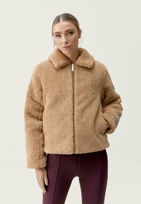 Kaha béžová fleecová bunda se zipem vpředu a rozšířeným límcem. Má strukturovaný povrch a široké rukávy, doplněné o těsně přiléhající legíny.