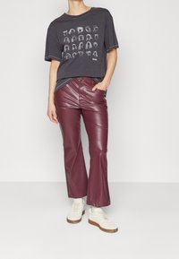 Person som bär en svart grafisk t-shirt med vita huvudskeppningar, burgundy faux läderbyxor, vita sneakers, står med en hand i fickan.