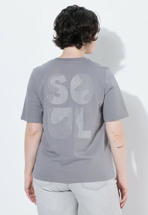 Personne aux cheveux courts et bouclés portant un t-shirt gris avec de grandes lettres stylisées dans le dos et un pantalon clair, de dos.