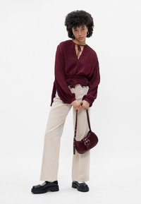 Blusa pregueada bordô com mangas longas, calças de perna larga bejes e uma pequena mala de ombro bordô. Sapatos plataforma pretos com sola robusta.