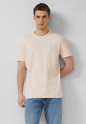 Młody mężczyzna z kręconymi włosami, ubrany w jasnoróżowy t-shirt z krótkim rękawem i niebieskie dżinsy, stoi na tle gładkiej szarej ściany.