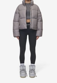 Person iført grå pufferjakke, sorte højtaljede tights, grå ankelhøje støvler og mærkevarer hvide sokker foran en hvid baggrund.