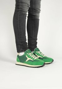 Zelené tenisky so zmesou semišových a syntetických materiálov, s béžovou medzipodrážkou a čiernou podrážkou, s detailne prepracovanými šnúrkami.