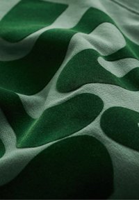 Zelená tkanina s různými abstraktními tmavě zelenými tvary, s texturovaným povrchem a hladkým pozadím, vytvářející vlnitý vzor.