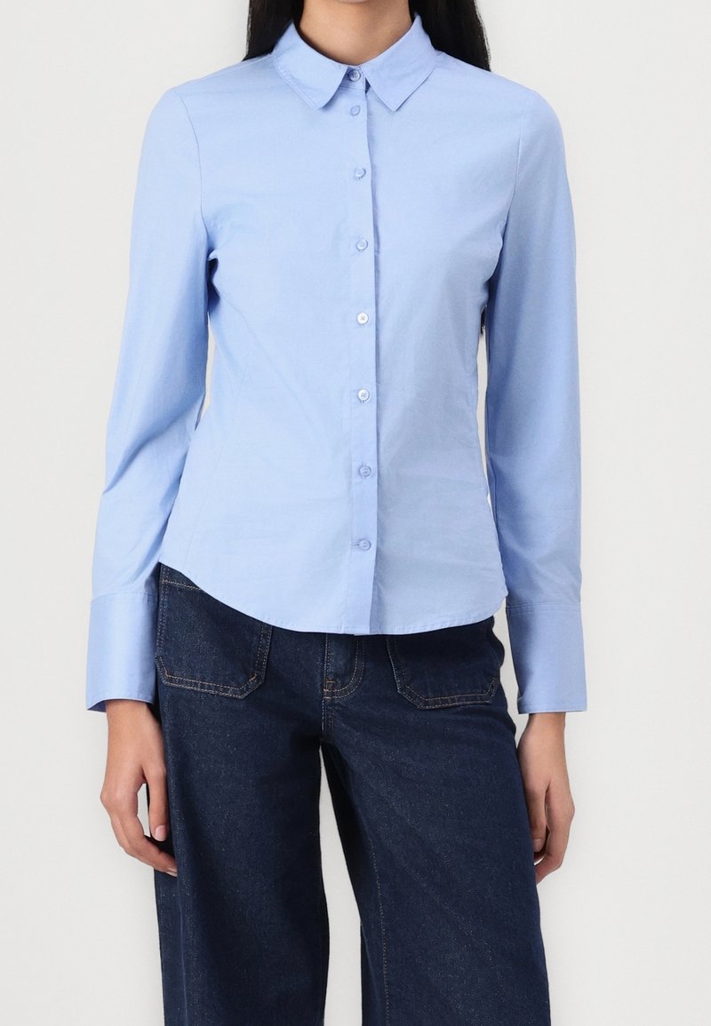 Femme portant une chemise bleu clair à boutons avec manches longues et un jean taille haute bleu foncé sur un fond uni.