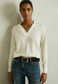 Cremfarbener V-Ausschnitt-Pullover mit strukturiertem Kragen, kombiniert mit blauen Jeans und einem braunen Gürtel mit goldener Schnalle. Überall glatte Textur.