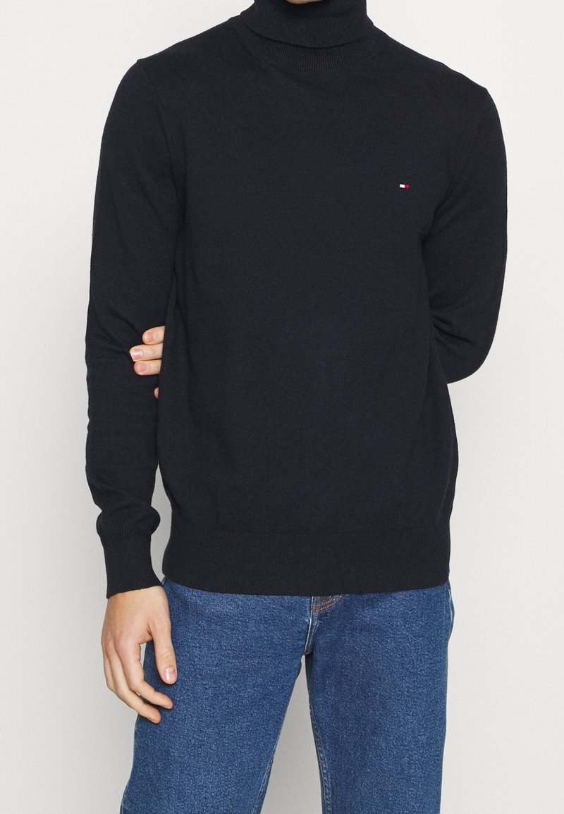 Uomo che indossa un maglione a collo alto nero con un piccolo logo sul petto e jeans blu, in piedi con un braccio dietro la schiena.