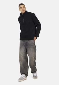 Pullover in pile nero con colletto alto e zip a metà, abbinato a jeans baggy grigi sbiaditi e scarpe da ginnastica grigie chiare.