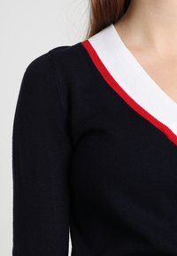 Suéter navy de manga larga, con un escote en V profundo y acentos de cuello acanalado en blanco y rojo que contrastan. Textura de punto suave.