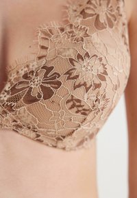 Spetsbehå i nude-färg med blommönster, genomskinlig textur och delikata avslutningsdetaljer längs kanterna.