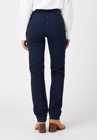 Moteris, vilkinti tamsiai mėlynus tiesius džinsus, baltą marškinių palaidinę su sagomis ir šviesiai rudais blokiniais auliniais batais, stovi atsisukusi atgal.