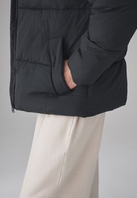 Giacca imbottita nera con chiusura a zip e tasca laterale, indossata sopra pantaloni beige chiaro, che mostra un tessuto strutturato.