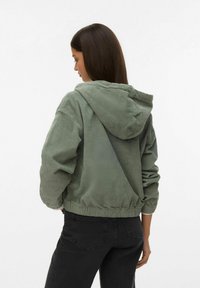 Overdimensioneret gr�øn fløjlstrøje med hætte, rynket bund og lange ærmer, parret med sorte jeans. Texturen virker blød og ribbet.