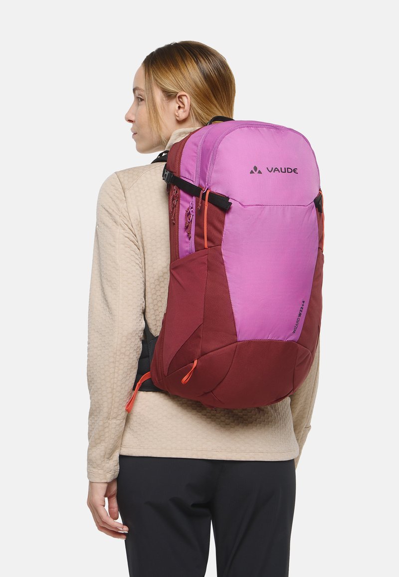 Mujer con una mochila Vaude bicolor en tonos rosa y vino, mirando hacia la izquierda, vestida con una chaqueta beige de textura y pantalones negros sobre un fondo blanco.