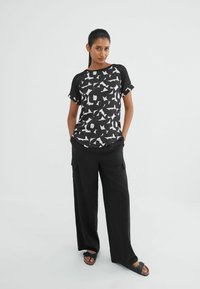 Kvinde iført sort og hvid abstrakt mønstret T-shirt, løse sorte cargo-bukser og sorte sandaler, står med hænderne i lommerne.