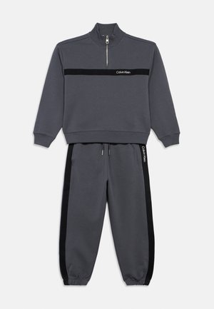 Un sweat gris avec un col montant, une demi-fermeture éclair et une bande noire sur la poitrine. Un jogging gris assorti présente des bandes latérales noires et un cordon de serrage.