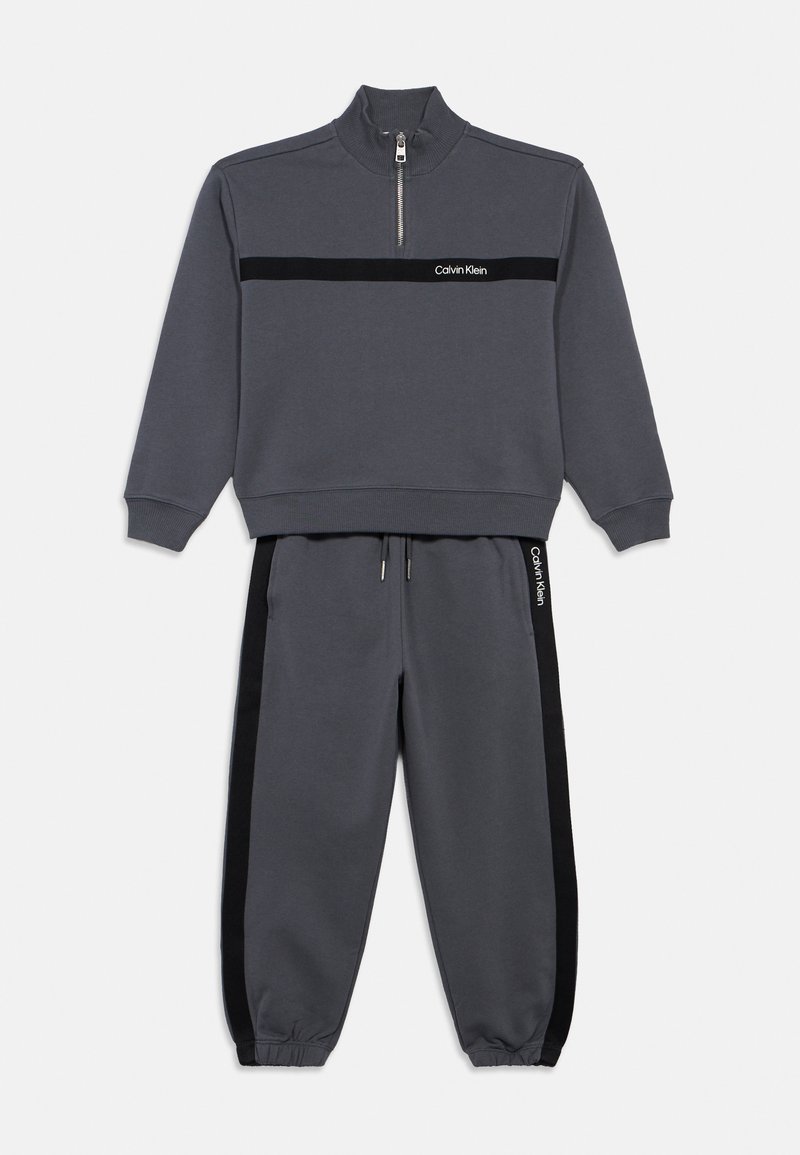 Szara bluza z półwysokim kołnierzem, zamek błyskawiczny do połowy oraz czarnym paskiem na piersiach. Pasujące szare dresy mają czarne paski po bokach oraz sznurek do regulacji.